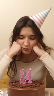 Üzgün bir kız elinde şenlikli bir pastayla bir masanın önünde oturuyor. 24 numaralı pastada mum sıkışıyor. Doğum günü kutlaması konsepti. dikey video