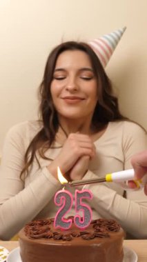 Kız elinde şenlikli bir pastayla bir masanın önünde oturuyor, içinde 25 numara şeklinde bir mum yanıyor ve söndürüyor. Doğum günü kutlaması kavramı. dikey video