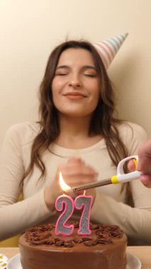 Kız elinde şenlikli bir pastayla bir masanın önünde oturuyor, 27 numara şeklinde bir mum yanıyor ve söndürüyor. Doğum günü kutlaması kavramı. dikey video