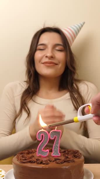 女の子はお祝いのケーキを持ってテーブルの前に座っており 27という数字の形をしたろうそくが燃えている 誕生日のお祝いコンセプト バーティカルビデオ — ストック動画