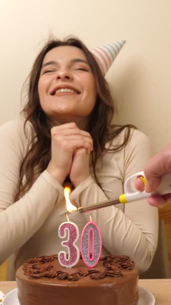 女の子は 30という数字の形をしたろうそくが燃えているお祝いのケーキでテーブルの前に座っており 彼女は吹き出します 誕生日のお祝いコンセプト バーティカルビデオ — ストック動画
