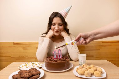 Masanın önünde elinde şenlikli bir pastayla oturan bir kız. 18 numaralı pastada bir mum yakılıyor. Doğum günü kutlaması konsepti.