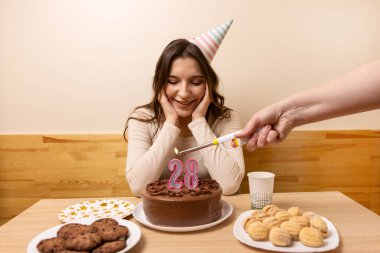 Bir kız elinde şenlikli bir pastayla bir masanın önünde oturuyor, üzerinde 28 numara şeklinde bir mum yanıyor. Doğum günü kutlaması konsepti.