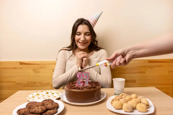 Bir kız elinde şenlikli bir pastayla bir masanın önünde oturur ve üzerinde 27 numara şeklinde bir mum söndürür. Doğum günü kutlaması konsepti.