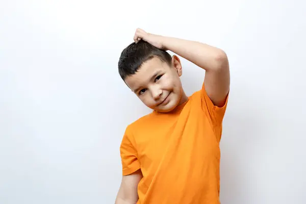 男の子がオレンジ色のTシャツで白い背景に立っている間に頭を掻いています ロイヤリティフリーのストック画像