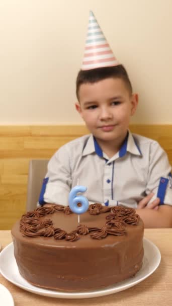 少年はお祝いのケーキを持ってテーブルの前に座っており そこでは6という数字の形をしたろうそくが燃えている 誕生日のお祝いコンセプト バーティカルビデオ — ストック動画