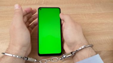 Kelepçeli bir adam ve yeşil ekranlı bir akıllı telefon. Akıllı telefon bağımlılığı kavramı.