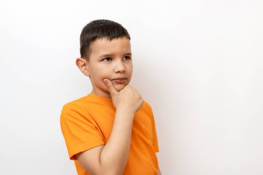 Beyaz arka planda turuncu tişörtlü bir çocuk elini çenesinde tutuyor..