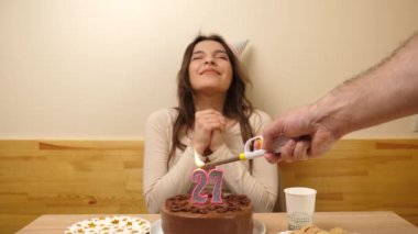 Kız elinde şenlikli bir pastayla bir masanın önünde oturuyor, 27 numara şeklinde bir mum yanıyor ve söndürüyor. Doğum günü kutlaması kavramı. Yavaş çekim