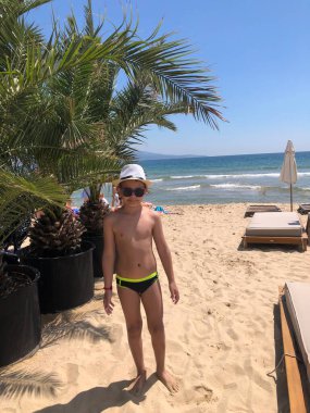 Şapkalı ve güneş gözlüklü bir çocuk, plajda palmiye ağaçlarının yanında poz veriyor..