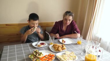 Anne ve oğlu masada oturmuş, yemek yiyor ve içki içiyorlar..