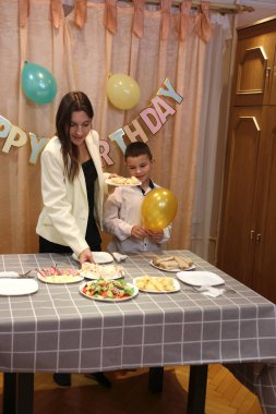 Bir anne, kızı ve oğluyla birlikte doğum günü kutlaması için şenlik masasına servis yapıyor. Yumuşak ışıklandırma.