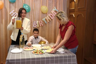 Bir anne, kızı ve oğluyla birlikte doğum günü kutlaması için şenlik masasına servis yapıyor. Yumuşak ışıklandırma.