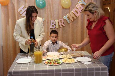 Bir anne, kızı ve oğluyla birlikte doğum günü kutlaması için şenlik masasına servis yapıyor. Yumuşak ışıklandırma.