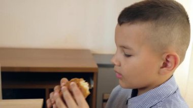 Çocuk masada otururken bir hamburger yiyor..