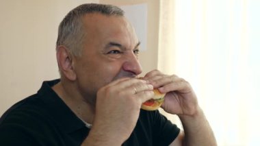 Bir adam masada otururken hamburger yiyor..