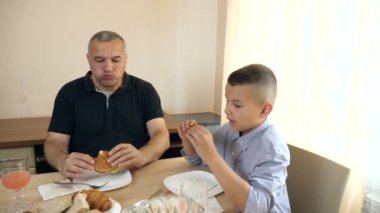 Baba ve oğul masada otururken hamburger yiyorlar..