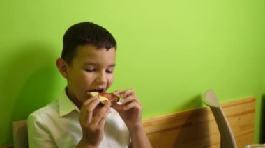 Çocuk masada otururken bir sandviç yiyor..