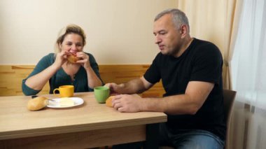 Bir erkek ve bir kadın mutfak masasında kahvaltı yapar, kahvelerini böreklerle içerler..