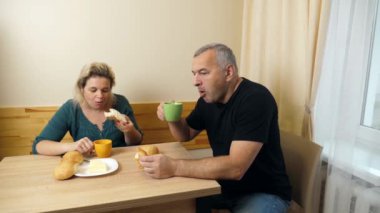 Bir erkek ve bir kadın mutfak masasında kahvaltı yapar, kahvelerini böreklerle içerler..