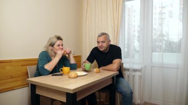 Bir erkek ve bir kadın mutfak masasında kahvaltı yapar, kahvelerini böreklerle içerler..