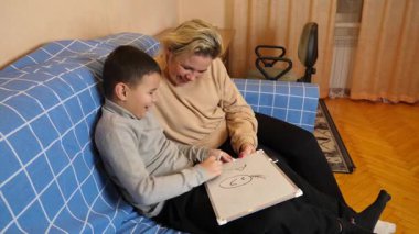 Anne ve oğul kanepede otururken tahta üzerine bir kalemle çizer..