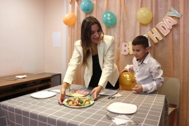 Bir anne, kızı ve oğluyla birlikte doğum günü kutlaması için şenlik masasına servis yapıyor. Yumuşak ışıklandırma.