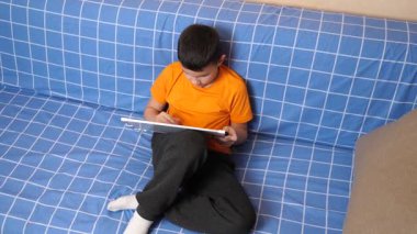 Bir çocuk kanepede otururken tahta üzerine bir kalemle resim çizer..