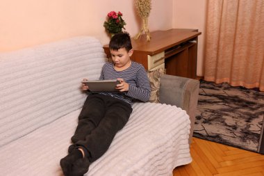 Bir çocuk kanepede otururken tablette oyun oynuyor..
