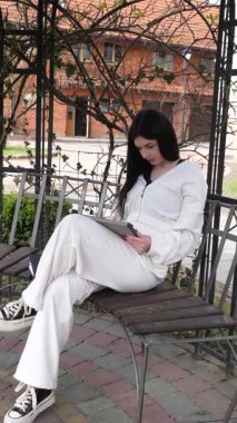 Bir kız öğrenci parkta bir bankta oturup tablet kullanarak ders çalışıyor ve notlar alıyor..