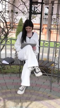 Bir kız öğrenci parkta bir bankta oturup tablet kullanarak ders çalışıyor ve notlar alıyor..