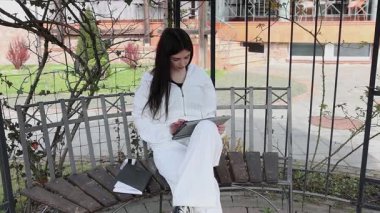 Bir kız öğrenci parkta bir bankta oturup tablet kullanarak ders çalışıyor ve notlar alıyor..