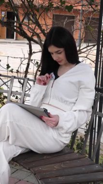 Bir kız öğrenci parkta bir bankta oturup tablet kullanarak ders çalışıyor ve notlar alıyor..