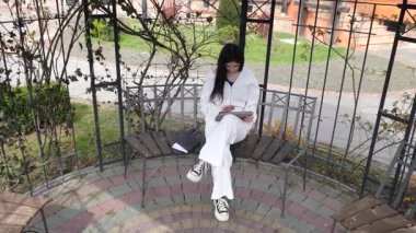 Bir kız öğrenci parkta bir bankta oturup tablet kullanarak ders çalışıyor ve notlar alıyor..