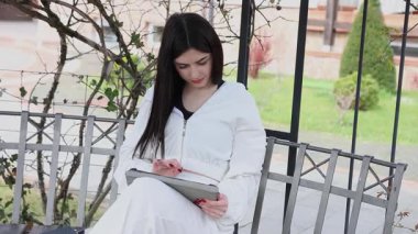 Bir kız öğrenci parkta bir bankta oturup tablet kullanarak ders çalışıyor ve notlar alıyor..