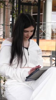 Bir kız öğrenci parkta bir bankta oturup tablet kullanarak ders çalışıyor ve notlar alıyor..