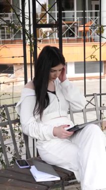 Bir kız öğrenci parkta bir bankta oturup tablet kullanarak ders çalışıyor ve notlar alıyor..