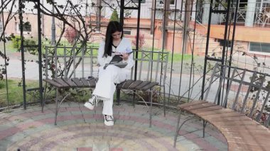 Bir kız öğrenci parkta bir bankta oturup tablet kullanarak ders çalışıyor ve notlar alıyor..