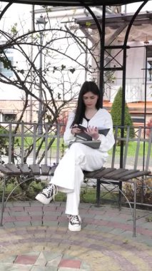 Bir kız öğrenci parkta bir bankta oturup tablet kullanarak ders çalışıyor ve notlar alıyor..