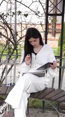 Bir kız öğrenci parkta bir bankta oturup tablet kullanarak ders çalışıyor ve notlar alıyor..