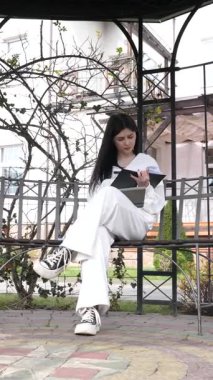 Bir kız öğrenci parkta bir bankta oturup tablet kullanarak ders çalışıyor ve notlar alıyor..