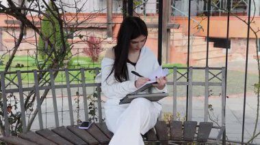 Bir kız öğrenci parkta bir bankta oturup tablet kullanarak ders çalışıyor ve notlar alıyor..