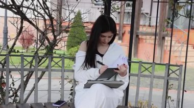 Bir kız öğrenci parkta bir bankta oturup tablet kullanarak ders çalışıyor ve notlar alıyor..