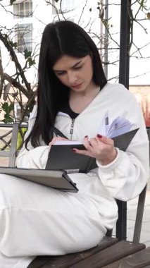 Bir kız öğrenci parkta bir bankta oturup tablet kullanarak ders çalışıyor ve notlar alıyor..