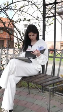 Bir kız öğrenci parkta bir bankta oturup tablet kullanarak ders çalışıyor ve notlar alıyor..