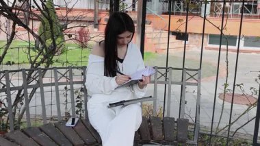 Bir kız öğrenci parkta bir bankta oturup tablet kullanarak ders çalışıyor ve notlar alıyor..