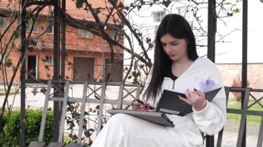 Bir kız öğrenci parkta bir bankta oturup tablet kullanarak ders çalışıyor ve notlar alıyor..