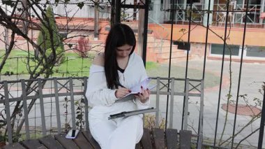 Bir kız öğrenci parkta bir bankta oturup tablet kullanarak ders çalışıyor ve notlar alıyor..