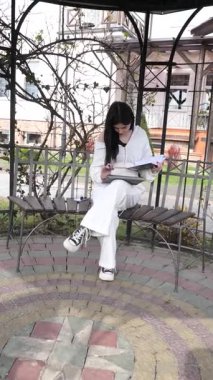 Bir kız öğrenci parkta bir bankta oturup tablet kullanarak ders çalışıyor ve notlar alıyor..