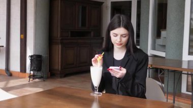 Bir kız sokakta masada otururken akıllı telefon kullanıyor ve kokteyl içiyor..